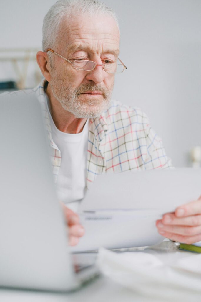 Elderly man with glasses reviews paperwork while sitting indoors with a laptop.